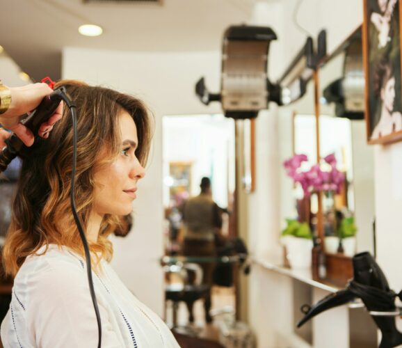 Hairdresser using curling tongs on customer's long brown hair in salon